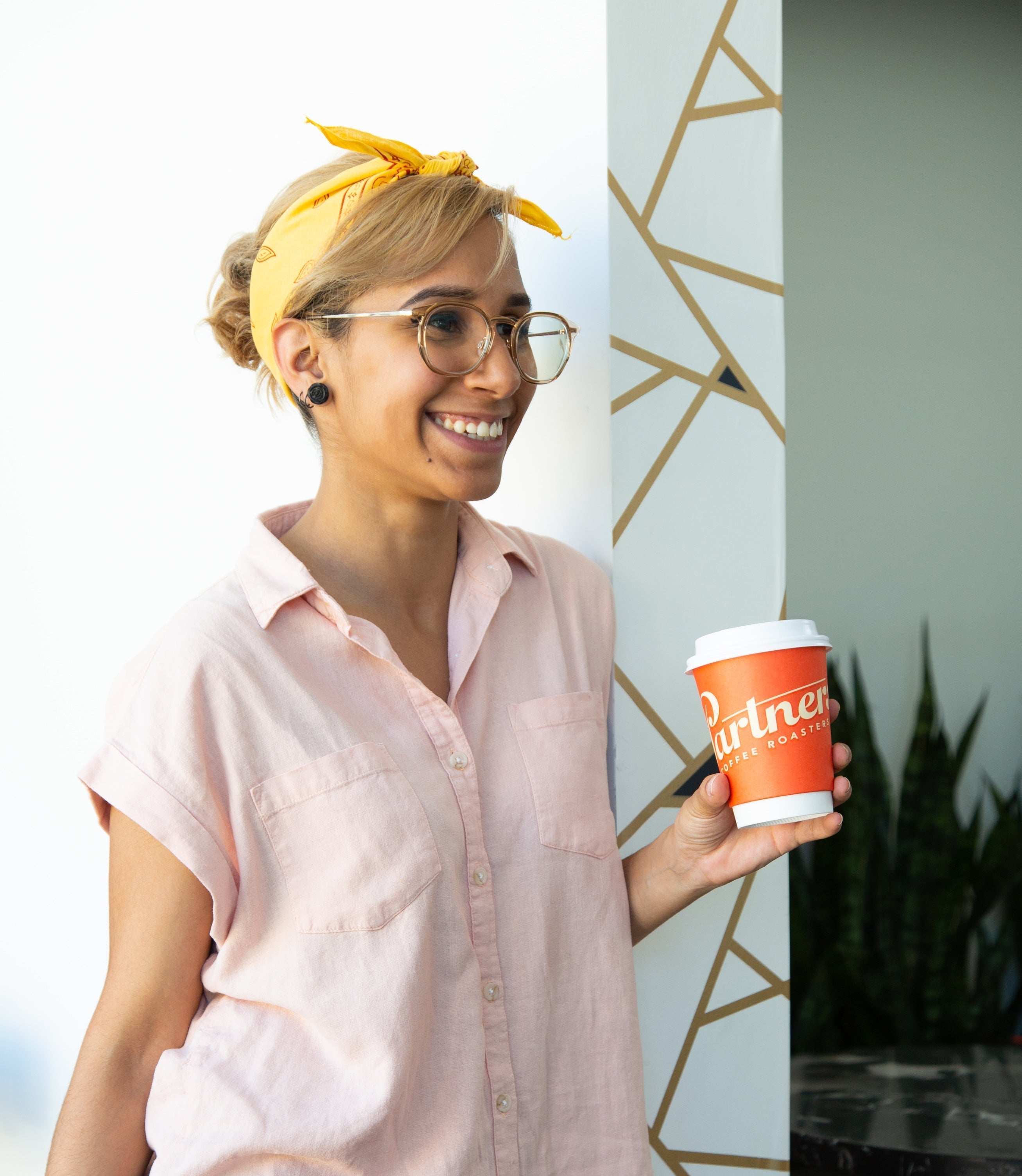 Person smiling and holding an orange Partners Coffee takeaway cup beside a geometric-patterned wall