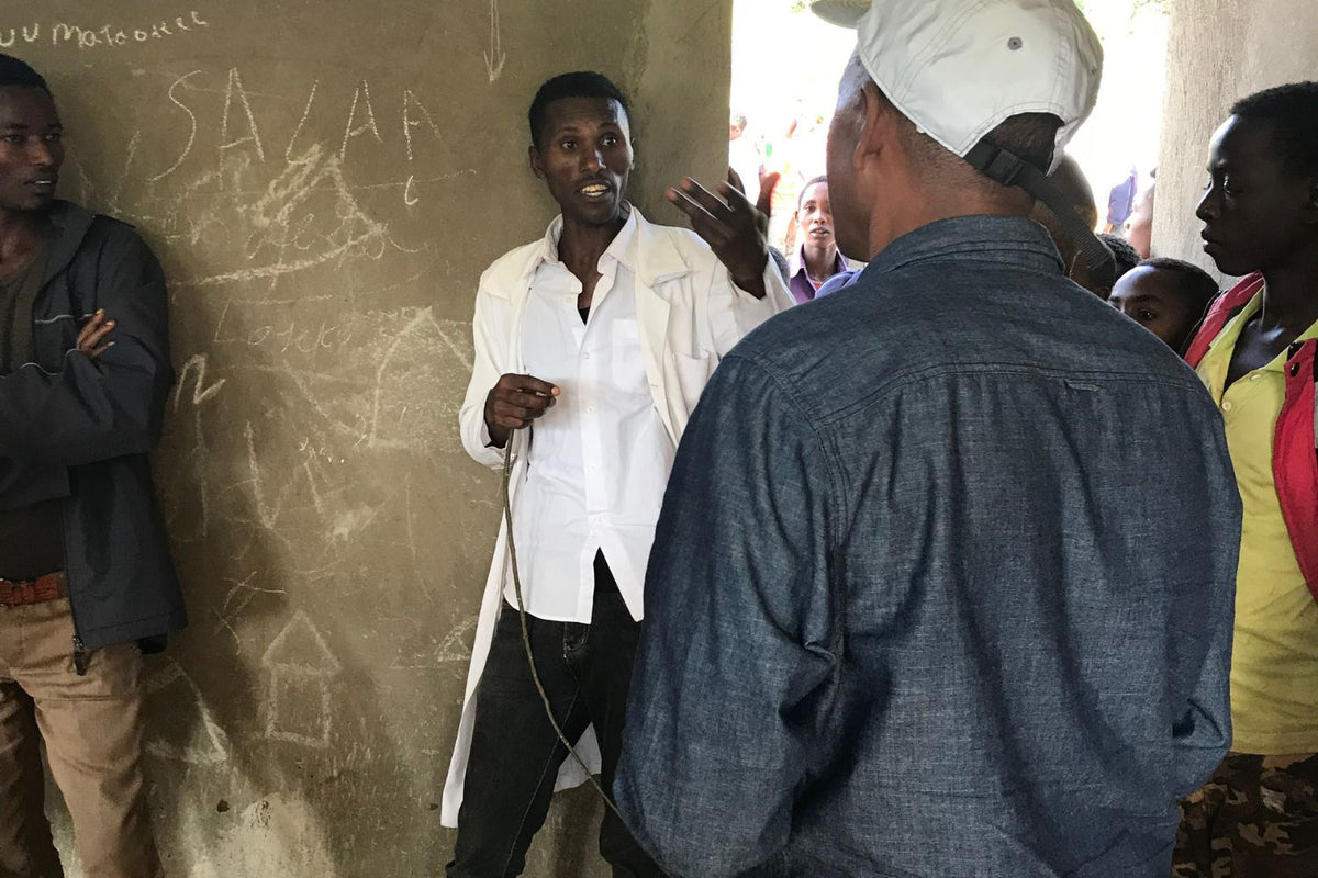 Man in a white coat speaking to a small group inside a concrete-walled room with chalk writing on the wall