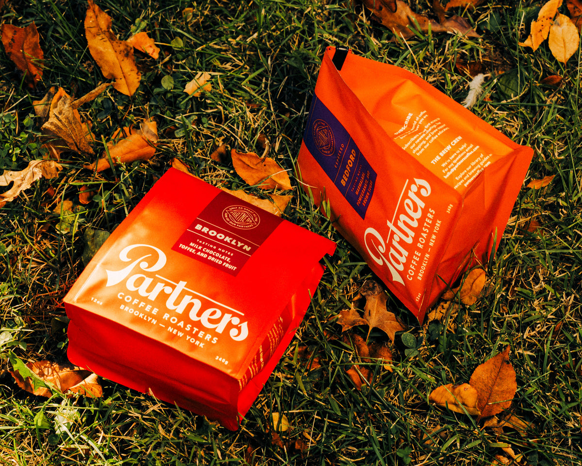 Two Partners Coffee bags on fallen leaves and grass, featuring Brooklyn and Colombia roasts