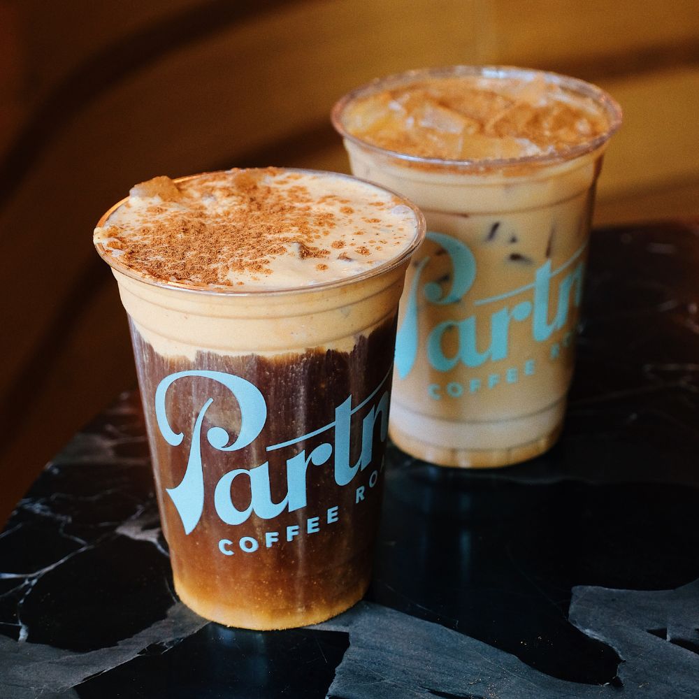 Three-quarter view of two iced coffee drinks with foamy, cinnamon-dusted tops on a dark marble table
