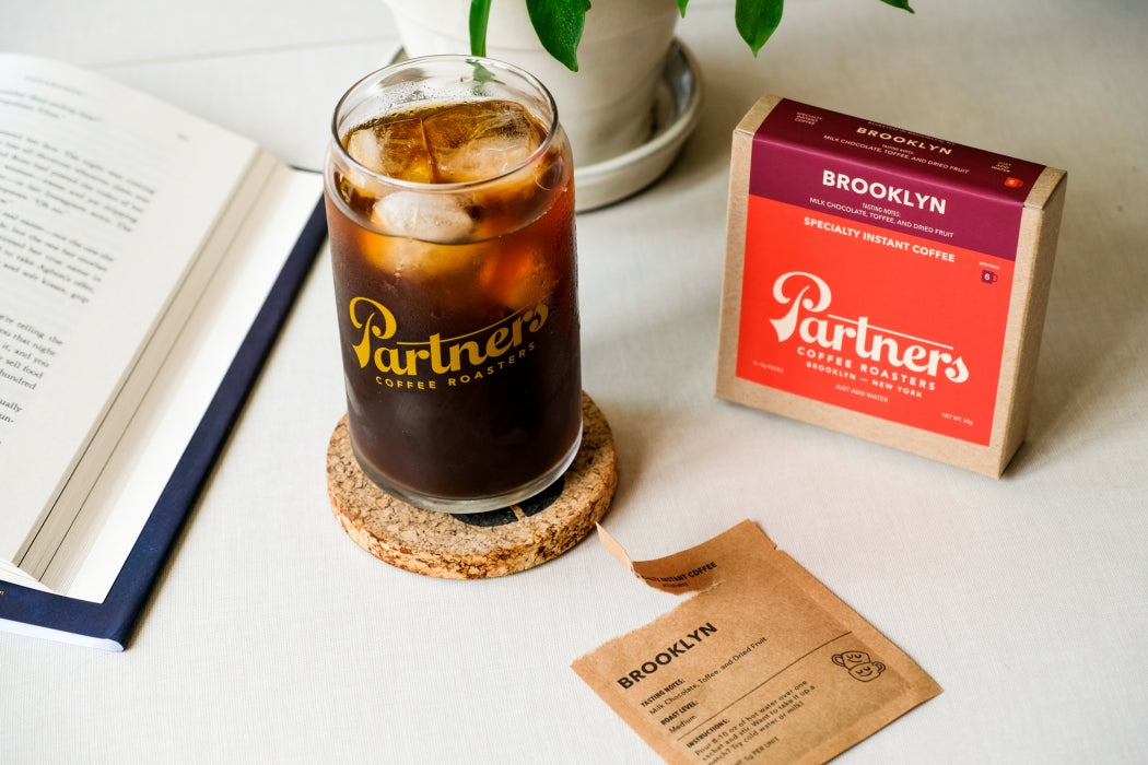 Angled view of Brooklyn specialty instant coffee box and Partners iced glass showcasing an opened sachet on a light tabletop