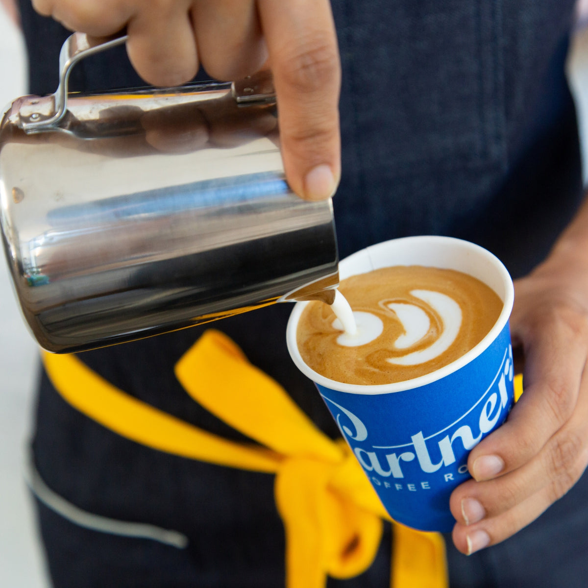Barista pouring steamed milk to create latte art in a blue Partners Coffee paper cup, close up of hands and stainless steel pitcher